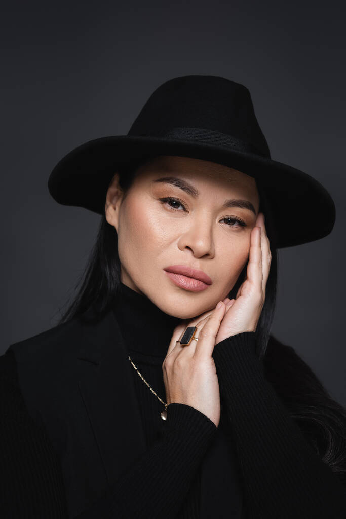 Porträt eines asiatischen Models in Blazer und Fedora-Hut, das isoliert auf dunkelgrau in die Kamera blickt - Foto, Bild