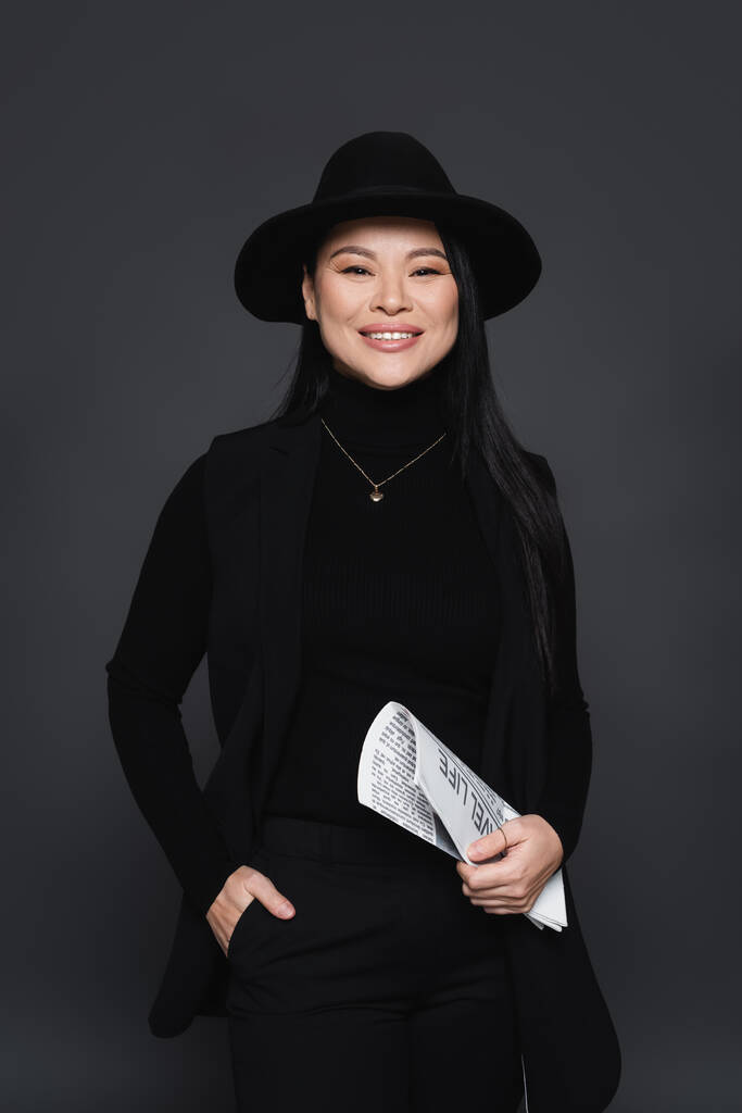 Usmívající se asijská žena v blejzru a fedora klobouk drží noviny izolované na tmavě šedé - Fotografie, Obrázek