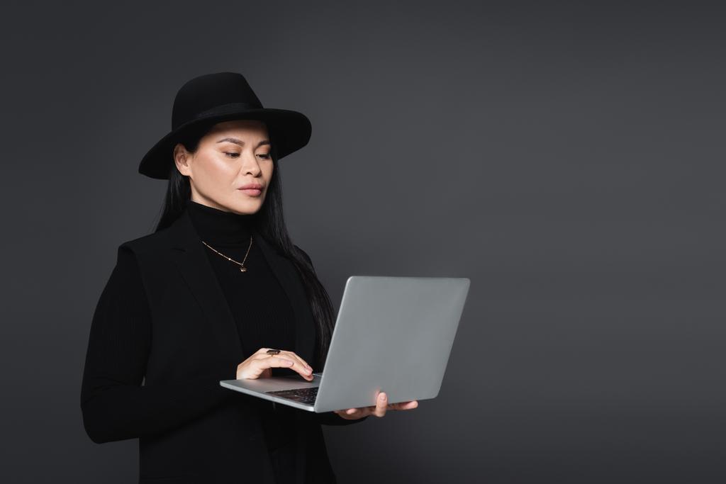 Stylish asian woman in fedora hat using laptop isolated on dark grey - Photo, Image