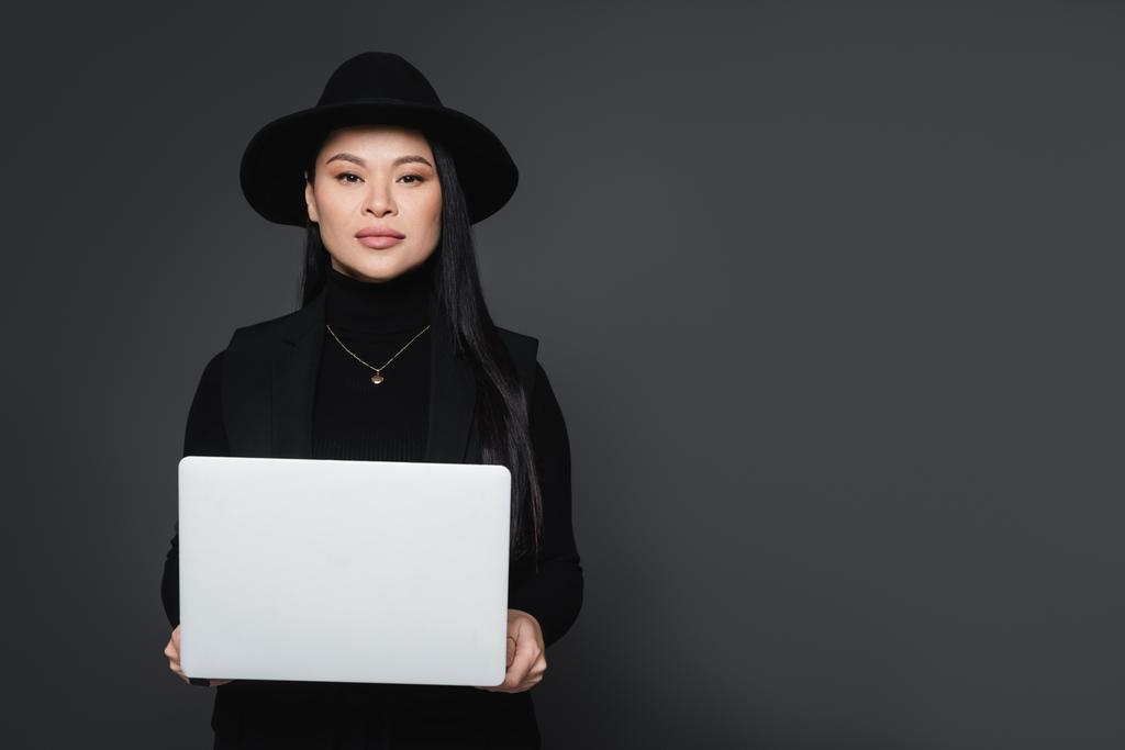 Stílusos ázsiai nő fedora kalap és garbó tartó laptop elszigetelt sötétszürke - Fotó, kép