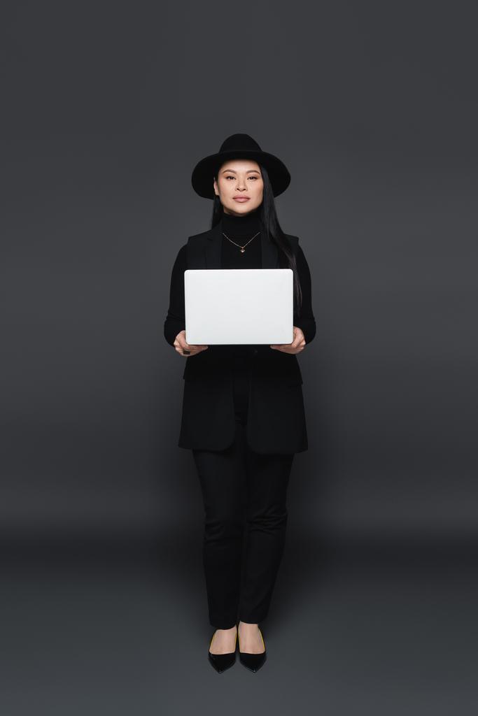 Longitud completa de la mujer asiática con estilo en sombrero fedora celebración portátil sobre fondo gris oscuro  - Foto, imagen