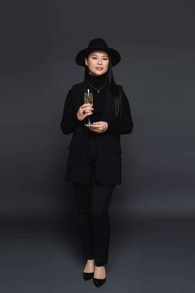 Longitud completa de elegante modelo asiático en sombrero fedora celebración de champán sobre fondo gris oscuro  - Foto, imagen