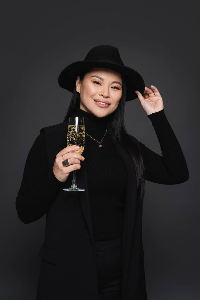 Lächelnde Asiatin mit Fedora-Hut und Blazer, die ein Glas Champagner isoliert auf dunkelgrau hält - Foto, Bild