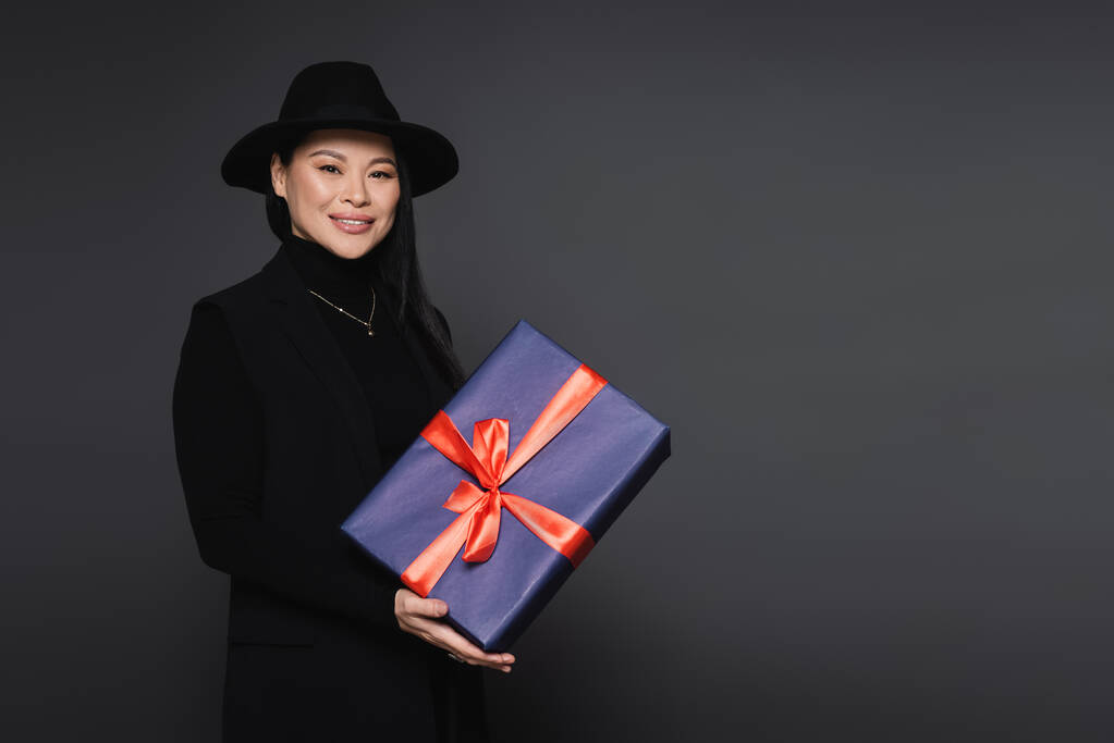 Lächelnde asiatische Frau mit Fedora-Hut hält Geschenk isoliert auf dunkelgrau - Foto, Bild