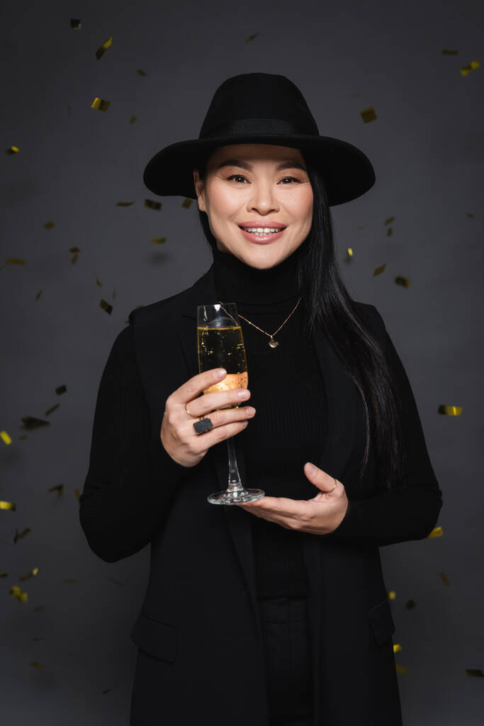 feliz asiático mujer en fedora sombrero celebración champán cerca confeti en oscuro gris fondo  - Foto, Imagen