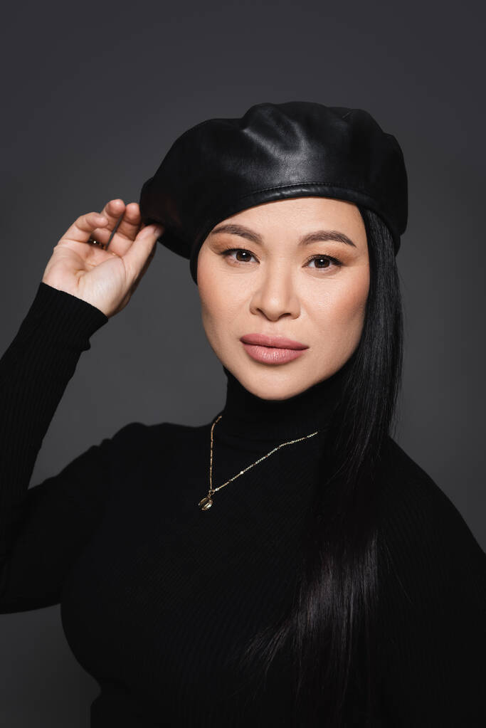 Asian model in beret and turtleneck looking at camera isolated on dark grey - Photo, Image