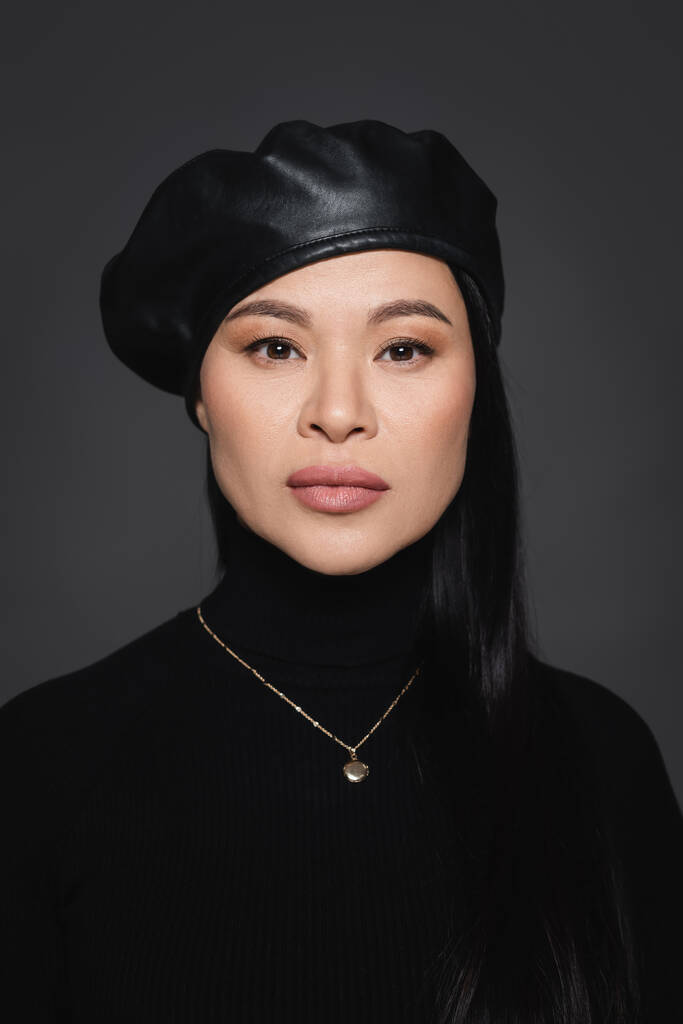Asiatisches Model mit Baskenmütze und Rollkragen blickt isoliert auf dunkelgraue Kamera - Foto, Bild