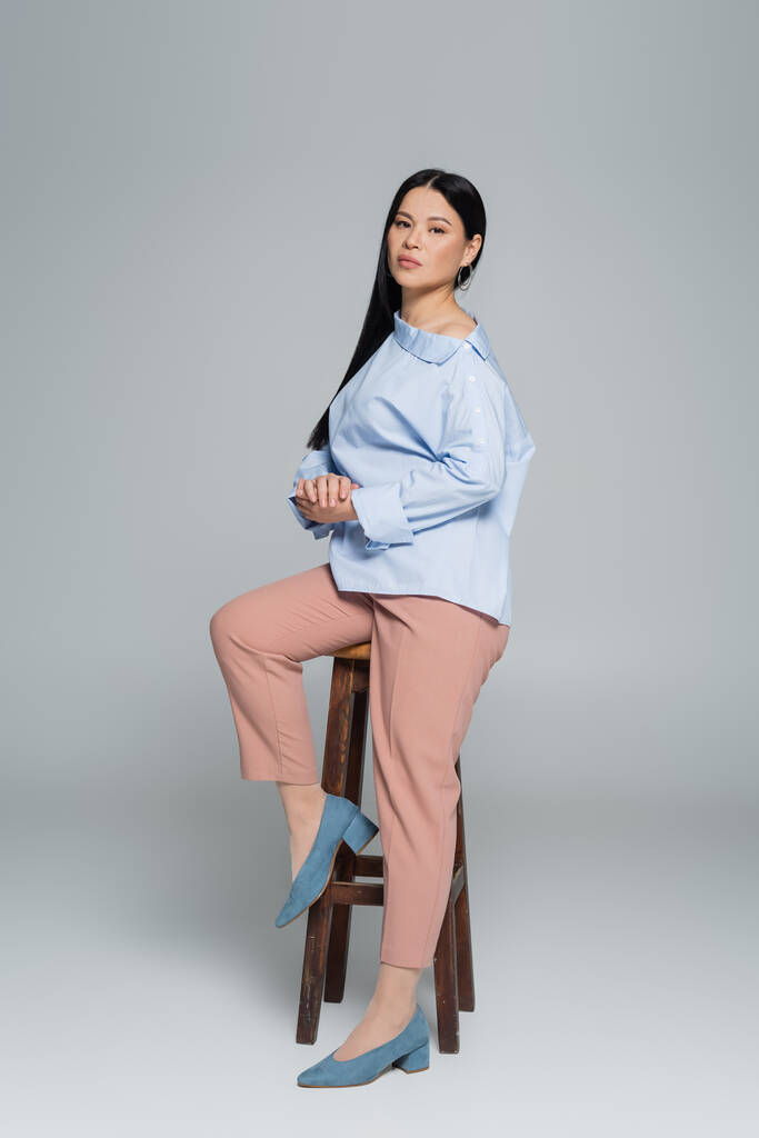 Longitud completa de elegante mujer asiática posando cerca de silla sobre fondo gris  - Foto, imagen