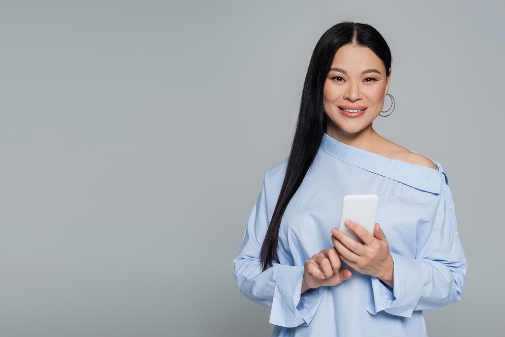 Positieve aziatische vrouw in blouse met behulp van mobiele telefoon geïsoleerd op grijs  - Foto, afbeelding