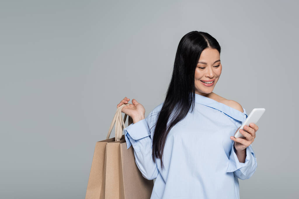 Akıllı telefon kullanan ve gri üzerinde alışveriş torbaları tutan pozitif Asyalı kadın.  - Fotoğraf, Görsel