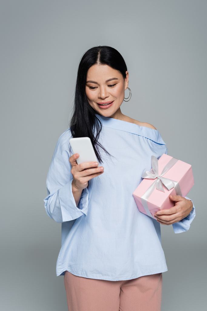 Позитивная азиатка, использующая смартфон и держащая подарок, изолированный на сером  - Фото, изображение