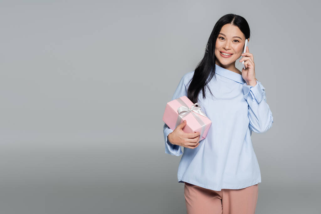 Veselá asijská žena drží dárek a mluví na smartphone izolované na šedé  - Fotografie, Obrázek