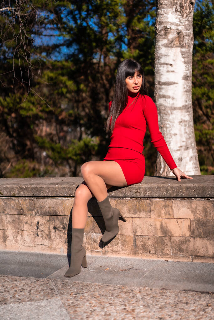 Jarní životní styl. Portrét mladé brunetky v parku s červenými šaty odpočívá a těší příroda sedí vedle stromu - Fotografie, Obrázek