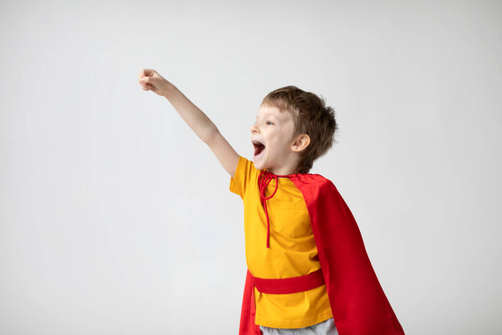 Menino sorridente em um traje de super-herói com um manto vermelho levantando a mão no fundo da parede branca. Conceito de felicidade e liberdade. - Foto, Imagem