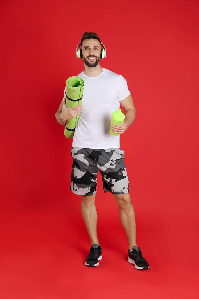 Przystojny mężczyzna ze słuchawkami, matą do jogi i shakerem na czerwonym tle - Zdjęcie, obraz
