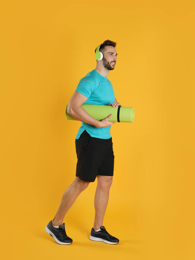 Knappe man met yogamat en hoofdtelefoon op gele achtergrond - Foto, afbeelding