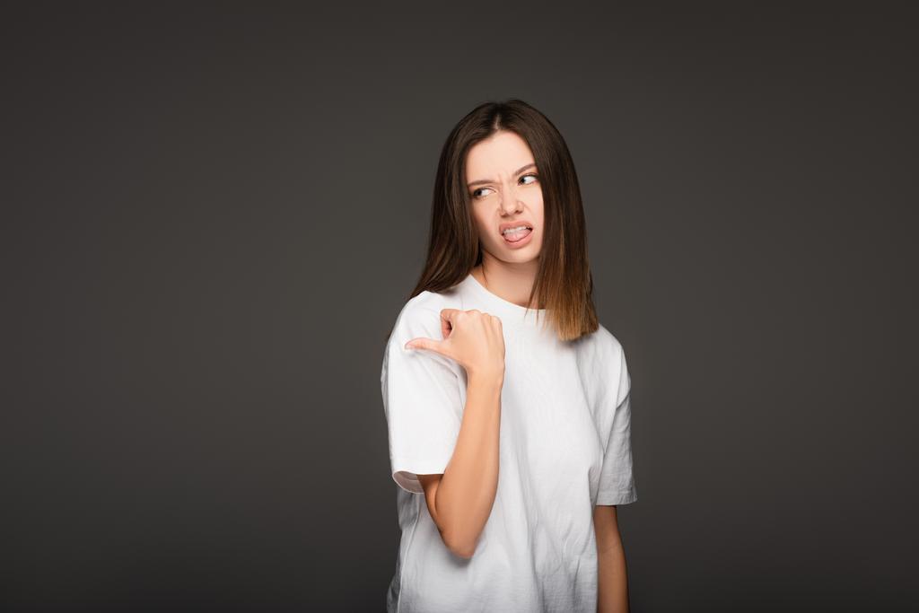Genç bir kadın kendini iğrenç hissediyor, dili dışarı çıkıyor ve parmağıyla işaret ediyor koyu gri renkte izole edilmiş. - Fotoğraf, Görsel