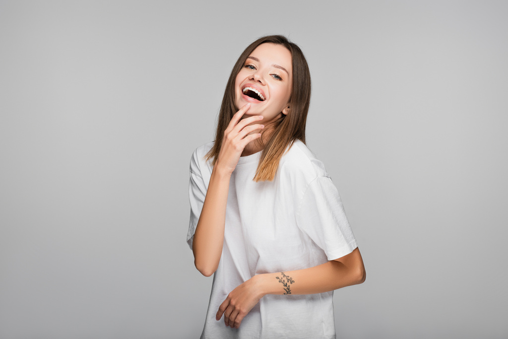 excited woman in white t-shirt laughing while looking at camera isolated on grey - Photo, Image