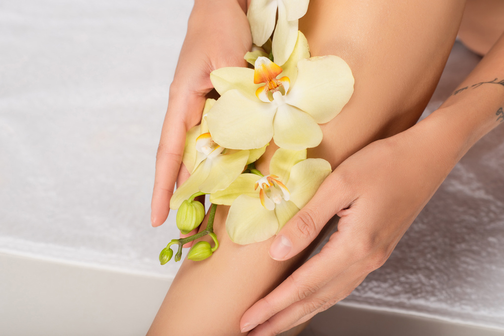 vista ritagliata di donna con orchidea su gamba liscia seduta su asciugamano bianco - Foto, immagini
