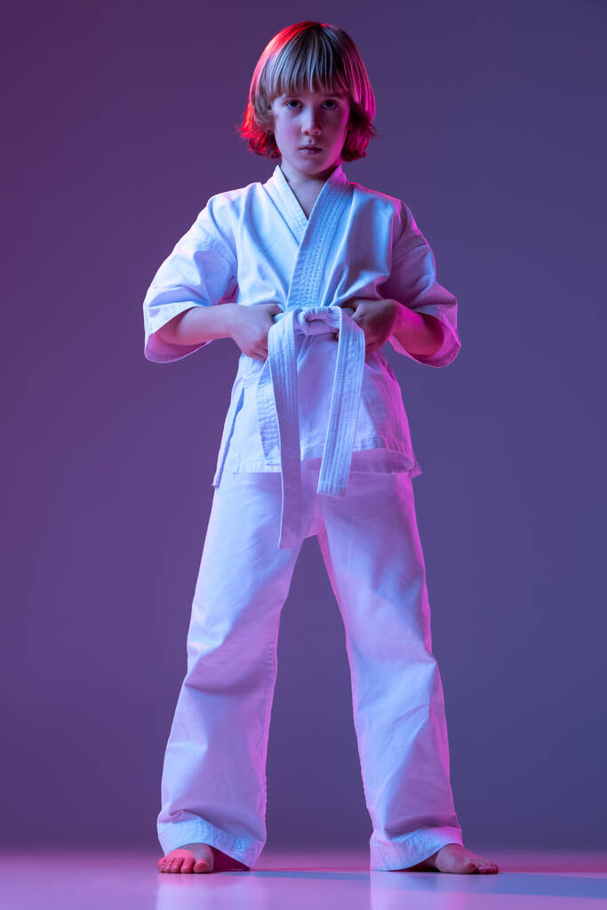 Enfant sérieux, petit garçon, athlètes de taekwondo ou de karaté en doboks posant isolés sur fond de couleur très péri en néon. Concept de sport, arts martiaux - Photo, image
