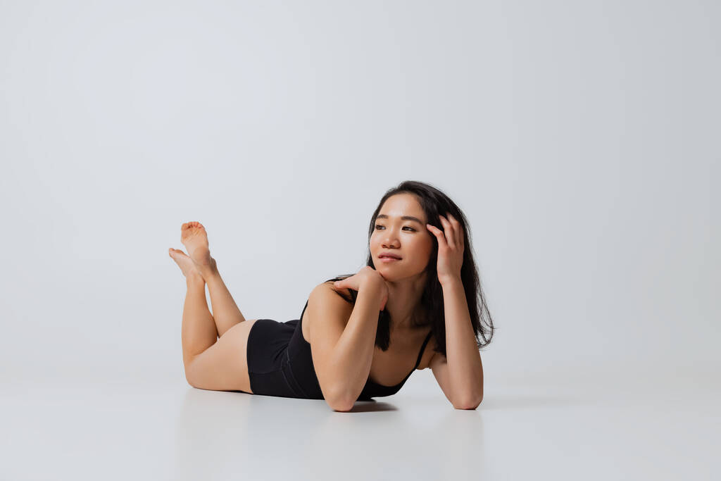 Portret pięknej Azjatki w czarnym body leżącej na podłodze, pozującej na szarym tle pracowni - Zdjęcie, obraz