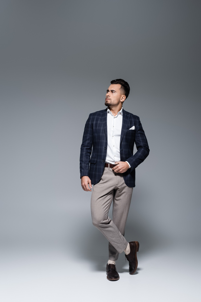 intera lunghezza del giovane uomo d'affari barbuto in giacca e cravatta in piedi con gambe incrociate su grigio - Foto, immagini