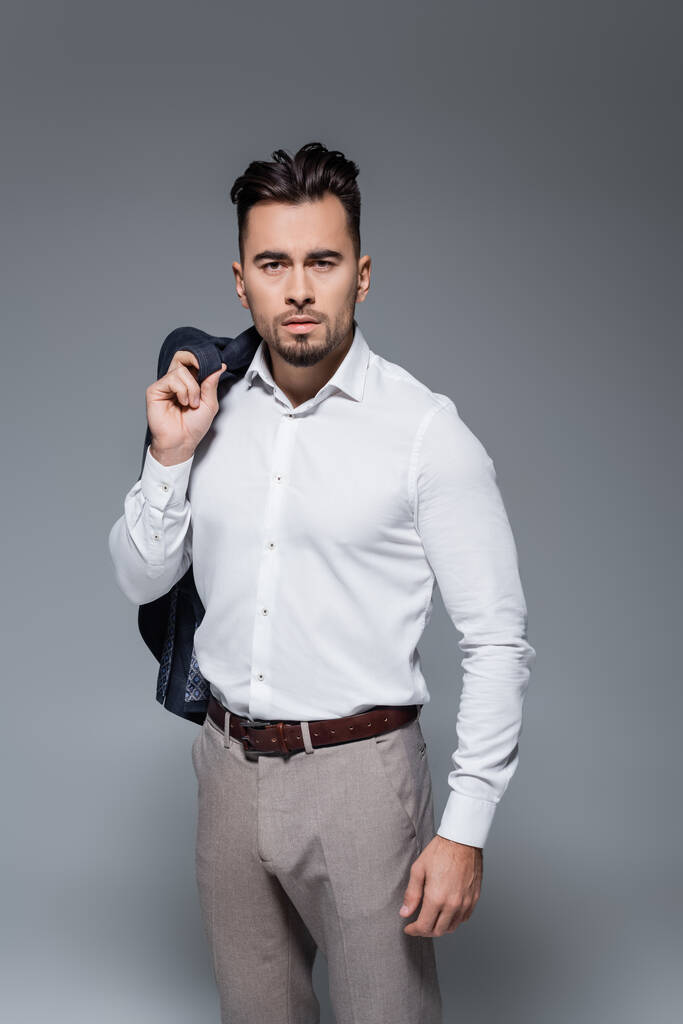 mladý vousatý podnikatel v bílé košili drží sako izolované na šedé - Fotografie, Obrázek