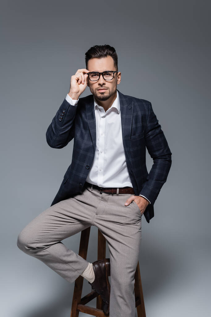 hombre de negocios barbudo en blazer ajustando gafas y sentado en la silla mientras posando con la mano en el bolsillo aislado en gris - Foto, imagen