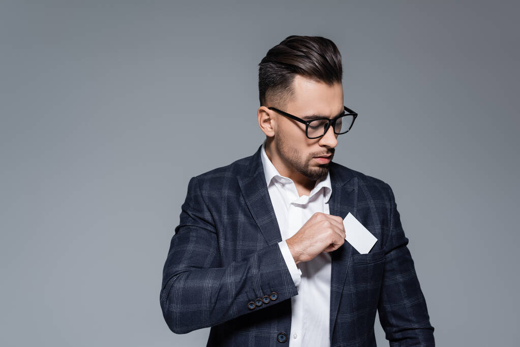 επιχειρηματίας με γυαλιά και σακάκι βάζοντας λευκή κάρτα στην τσέπη απομονωμένη σε γκρι - Φωτογραφία, εικόνα