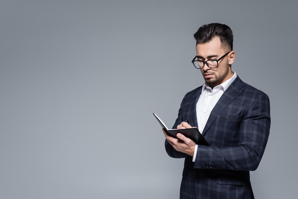 mladý podnikatel v obleku a brýle psaní v notebooku izolované na šedé - Fotografie, Obrázek