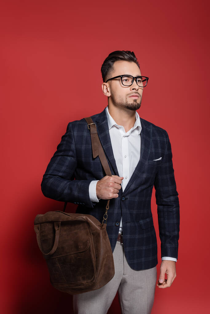 homem de negócios barbudo em blazer e óculos de pé com saco de couro em vermelho - Foto, Imagem