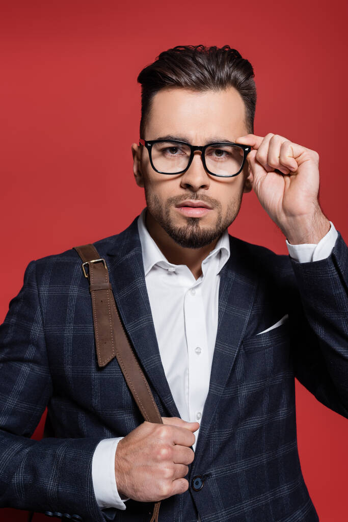 vousatý podnikatel v blejzru nastavuje brýle a drží koženou tašku na červené - Fotografie, Obrázek