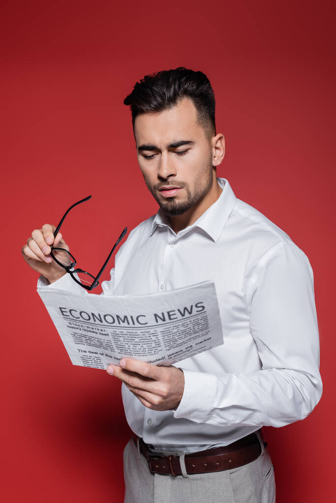 hombre de negocios barbudo con camisa blanca sosteniendo gafas y leyendo noticias sobre rojo - Foto, Imagen