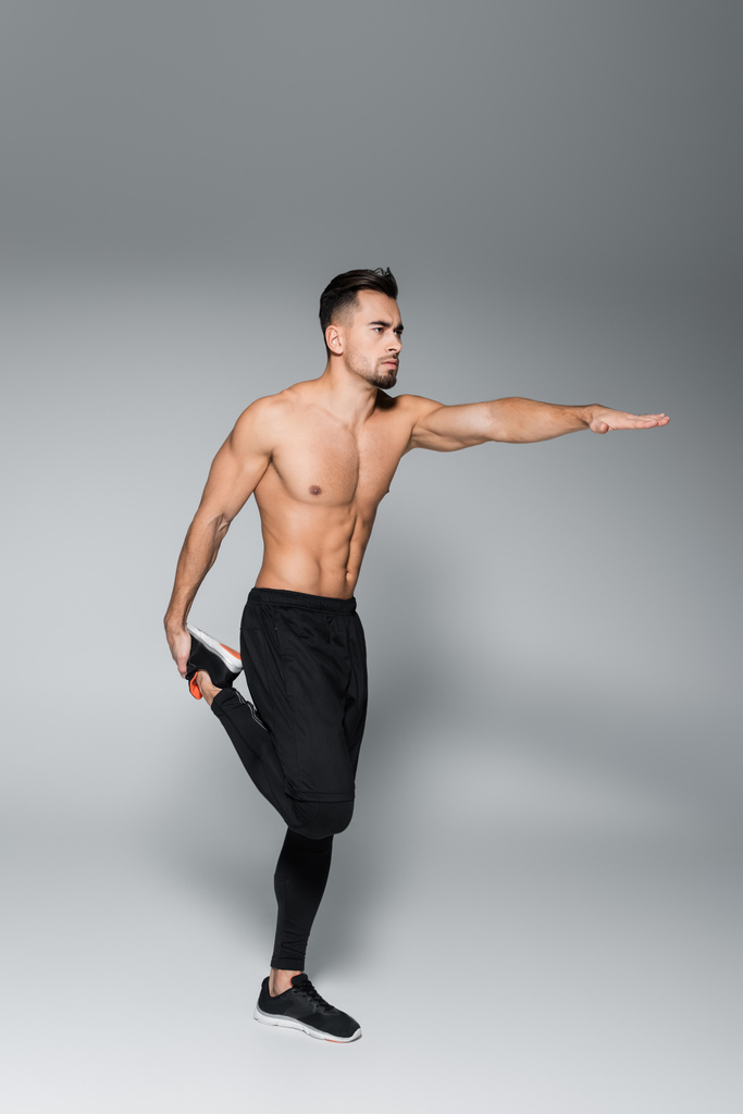 volledige lengte van de jonge shirtloze sportman stretching leg en staan met uitgestrekte hand op grijs - Foto, afbeelding
