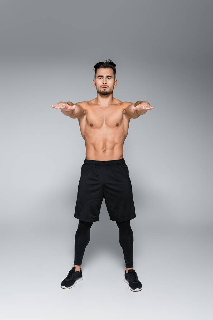 full length of young shirtless sportsman exercising with outstretched hands on grey - Photo, Image