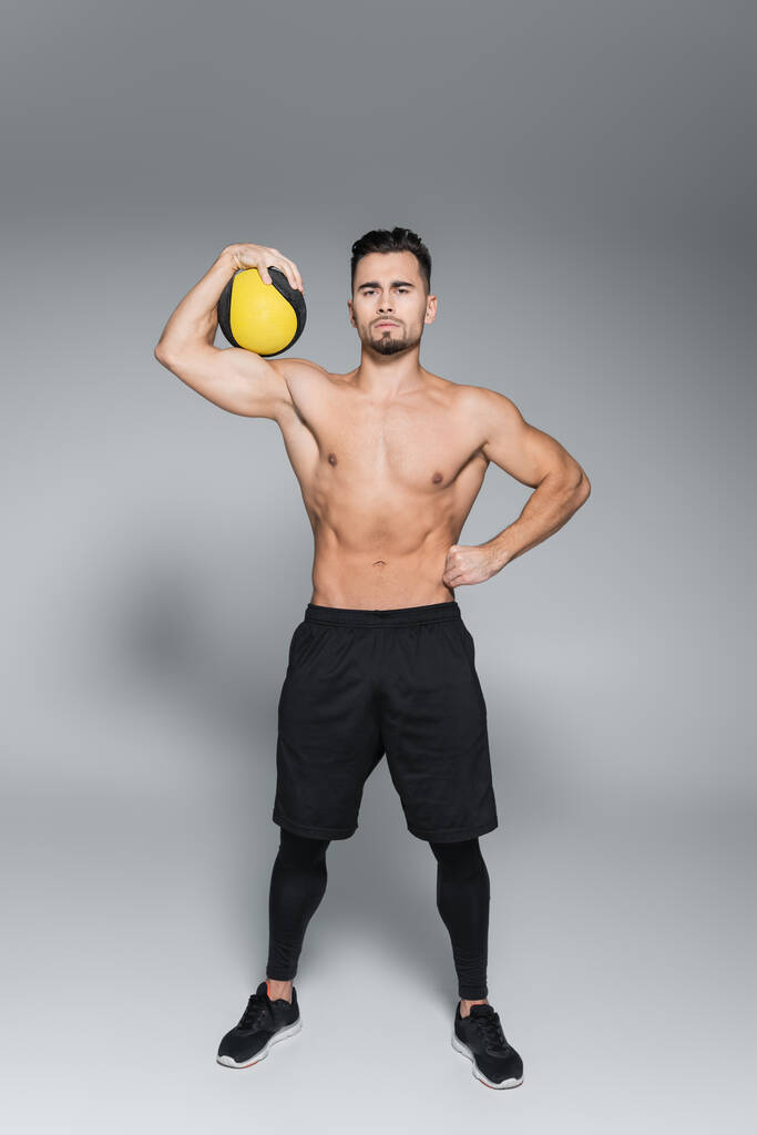 volledige lengte van shirtloze en bebaarde sportman die bal vasthoudt en met de hand op de heup op grijs staat - Foto, afbeelding