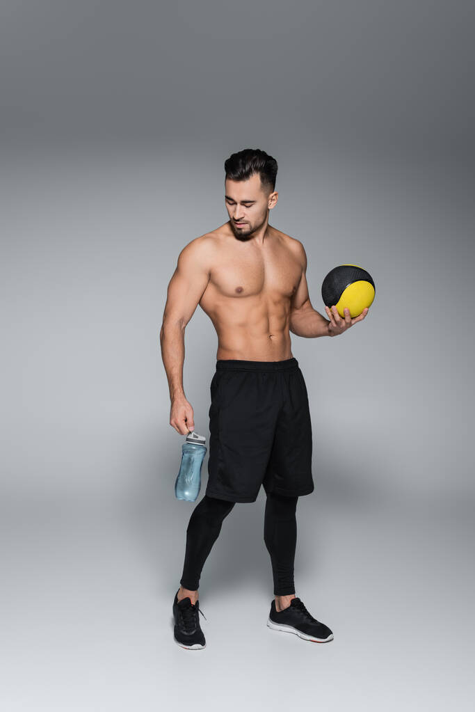 comprimento total do desportista sem camisa e barbudo segurando bola e garrafa de esportes em cinza - Foto, Imagem