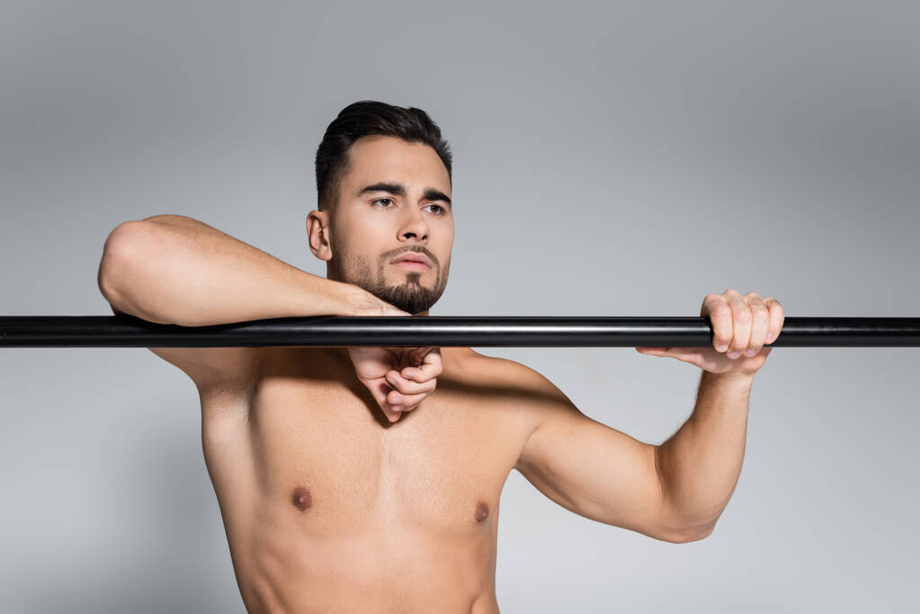 deportista musculoso mirando hacia otro lado y apoyándose en la barra horizontal aislado en gris - Foto, Imagen