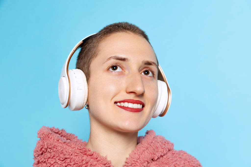 Glückliches hübsches Mädchen, Studentin mit kurzen Haaren, rosa Kapuzenpullover mit Kopfhörern auf blauem Hintergrund. Konzept von Emotionen, Musik - Foto, Bild