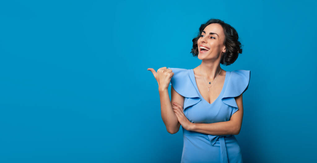 Photo en gros plan d'une belle jeune femme heureuse en robe bleue, qui pointe du doigt et isolée sur fond bleu - Photo, image