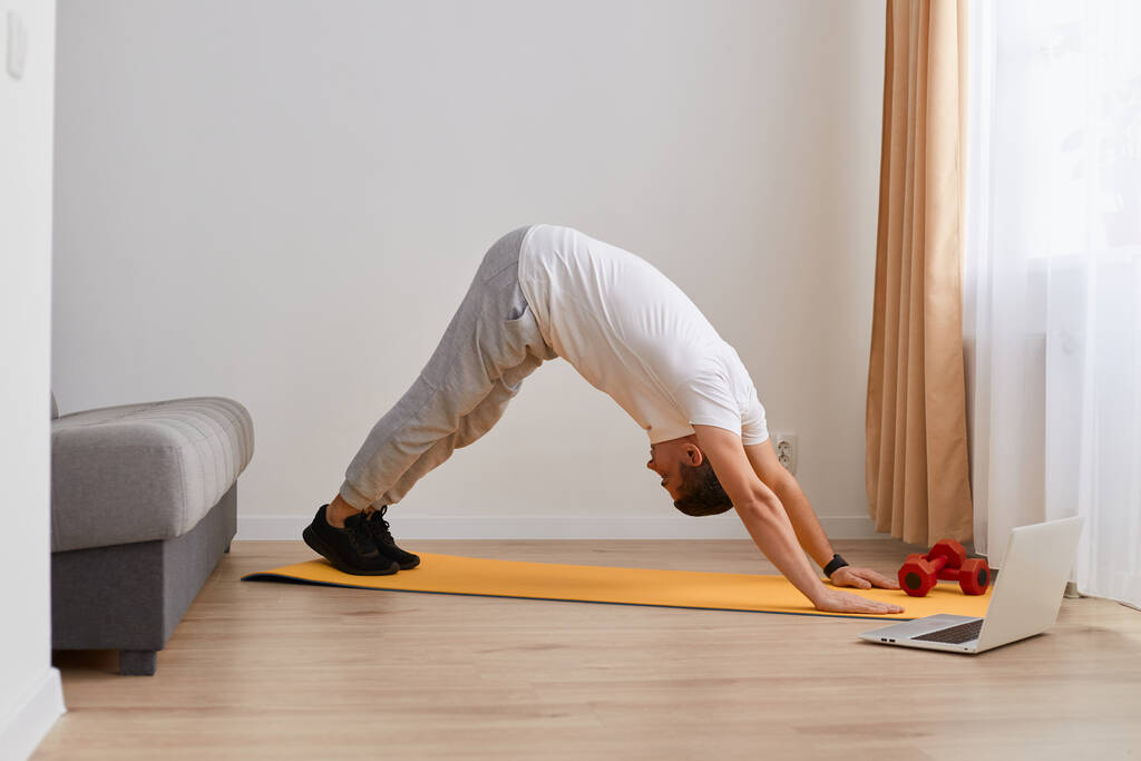 Retrato de un joven adulto caucásico deportista practicando yoga, haciendo ejercicio de perro orientado hacia abajo, pose adho mukha svanasana, haciendo ejercicio, usando ropa deportiva ligera. - Foto, imagen