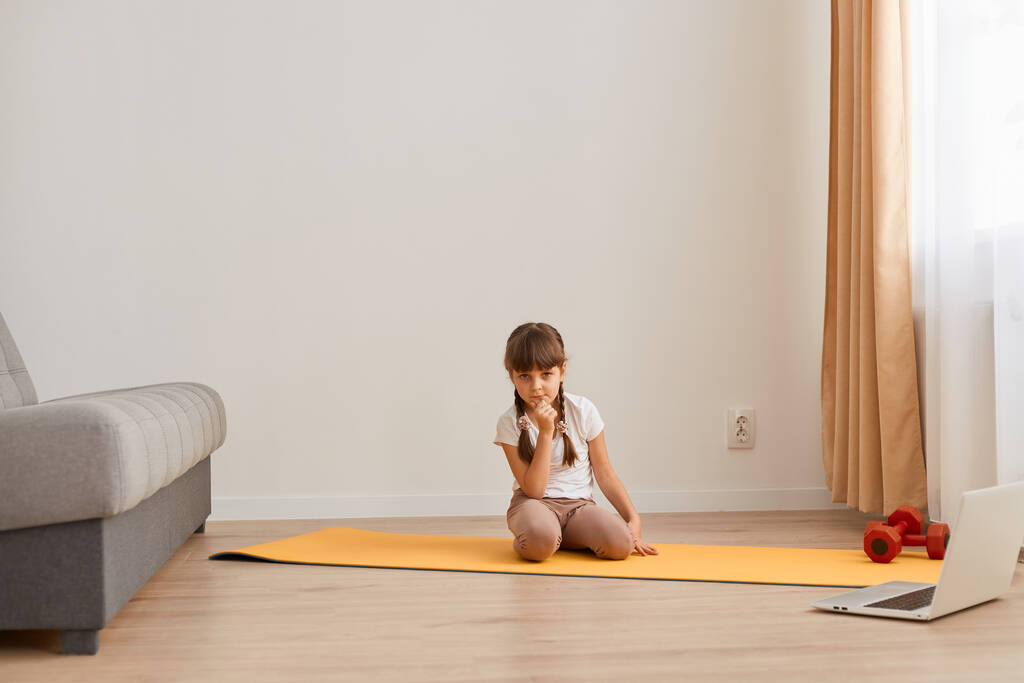 Εσωτερική λήψη του μικρού κοριτσιού που κάθεται σε ένα χαλάκι στο πάτωμα και κοιτάζει την κάμερα, να ξεκουράζεται ενώ κάνει αθλητικές ασκήσεις στο σπίτι μόνη της, παιδική φροντίδα υγείας και φυσικής κατάστασης. - Φωτογραφία, εικόνα