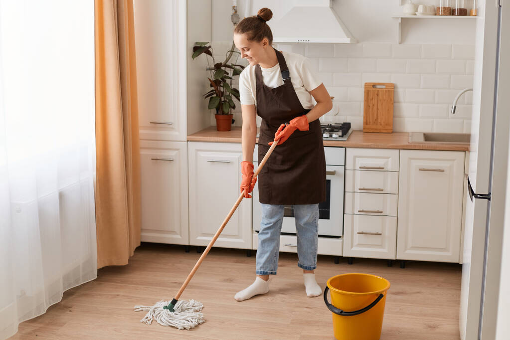Portret van een vrouwelijke huishoudster met dweil en emmer met schoonmaakmiddel, huisvrouw met spijkerbroek en bruin schort, schoonmaken van haar keuken. - Foto, afbeelding