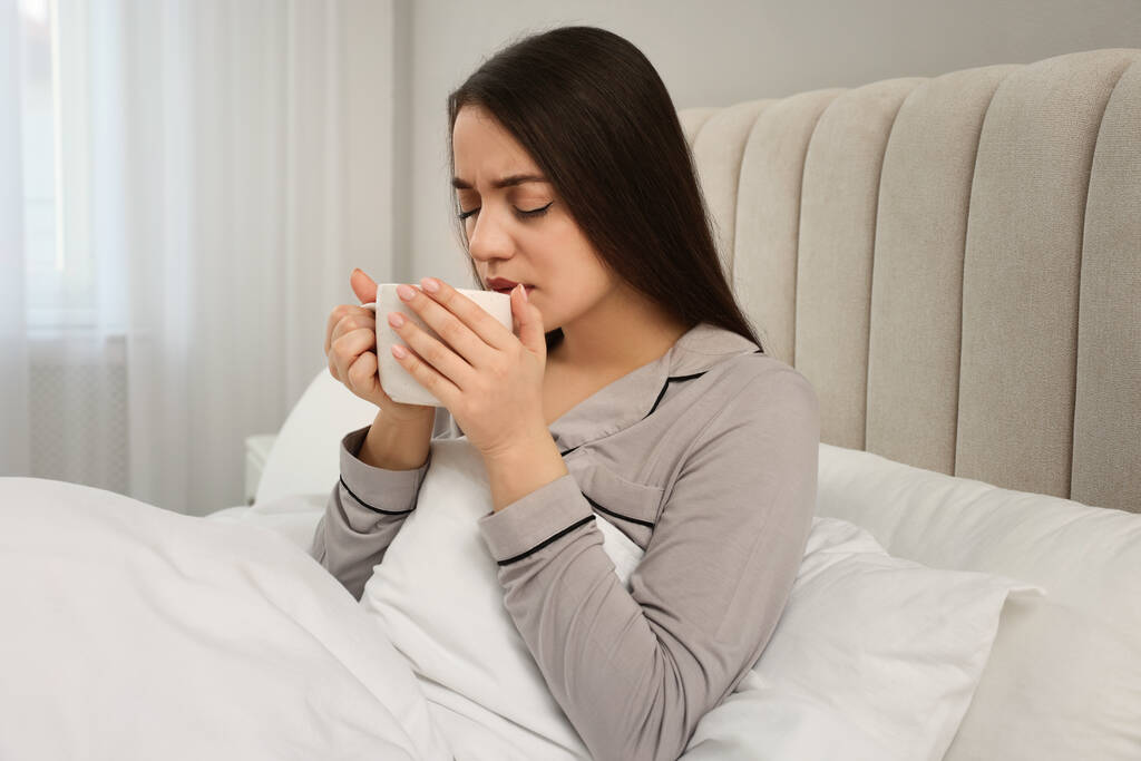 Άρρωστη νεαρή γυναίκα με φλιτζάνι ζεστό ρόφημα στο κρεβάτι στο σπίτι - Φωτογραφία, εικόνα
