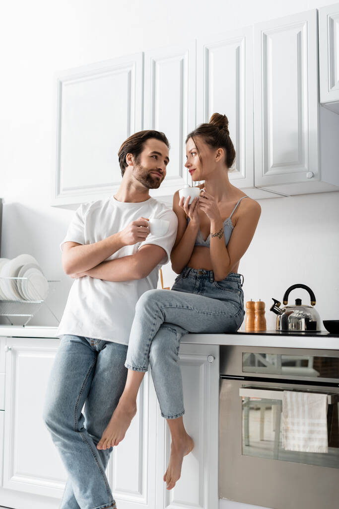 mulher alegre com tatuagem segurando xícara de café e sentado perto do namorado na cozinha  - Foto, Imagem