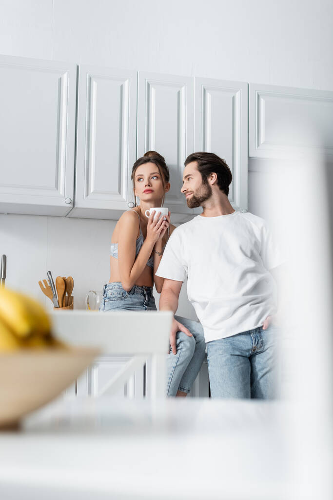 tattooed young woman in bra holding cup near bearded boyfriend in kitchen  - Photo, Image