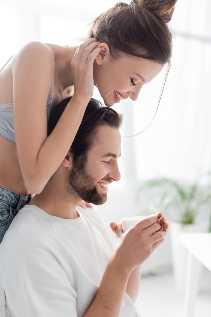 vista lateral da mulher alegre em sutiã de seda de pé perto namorado com copo  - Foto, Imagem