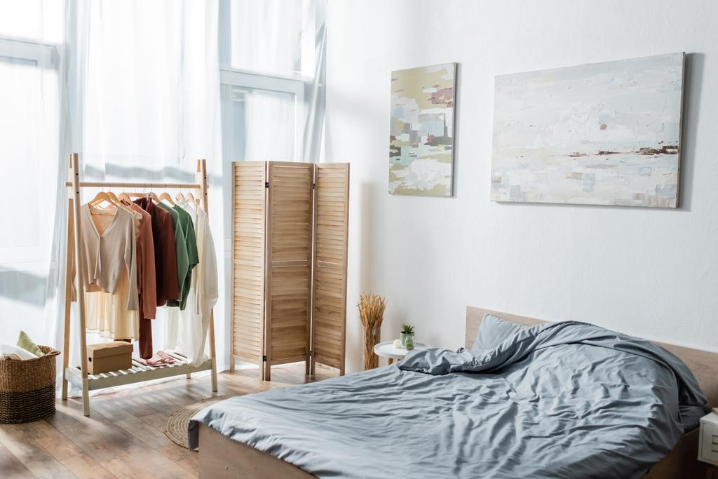 cama moderna cerca de la pantalla plegable y rack con ropa en el dormitorio  - Foto, imagen