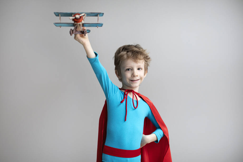 Il piccolo supereroe con un mantello rosso gioca con un aeroplano. Buon bambino sorridente. Successo, motivazione e concetto di viaggio. - Foto, immagini