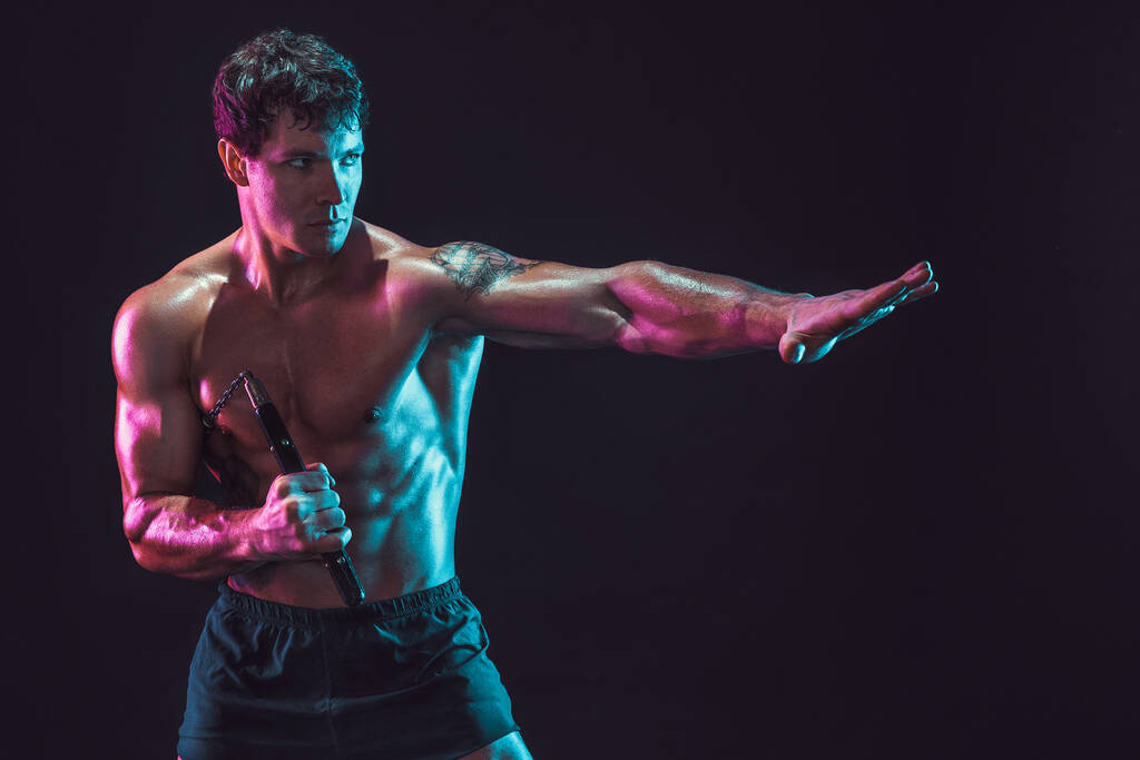 黒を背景に隔離されながらヌンチャクで修行する筋肉の男の肖像。スポーツコンセプト - 写真・画像